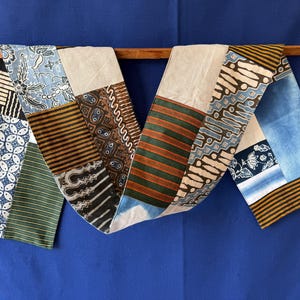 Peut inclure: Un long chemin de table en tissu présentant un motif patchwork composé de divers imprimés batik. Le textile affiche un mélange de couleurs bleu, marron, vert et crème avec des motifs géométriques, floraux et rayés en panneaux verticaux.
