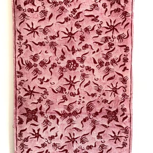 May include: A pink fabric with a red batik pattern. The pattern features a repeating design of stars, flowers, and swirls.