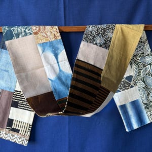 Peut inclure: Une longue écharpe patchwork rectangulaire composée de divers panneaux de tissu dans des tons de bleu, marron, beige, noir et blanc. Le textile présente des motifs variés incluant des rayures, des fleurs, des formes géométriques et des effets tie-dye.