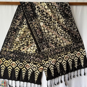 Könnte beinhalten: Ein rechteckiges Textiltuch mit schwarzem Grund und komplexen Batik-Mustern in Creme, Braun und Beige. Das Design umfasst geometrische Rautenformen und wiederkehrende florale Motive, abgeschlossen mit Fransen an der unteren Kante.