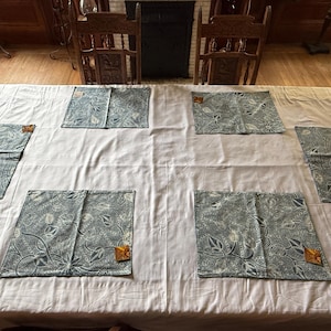May include: A large, light gray rectangular tablecloth featuring six rectangular placemats arranged in a grid. Each placemat displays a dark blue and white botanical pattern with intricate leaf and vine motifs and a small brown tag attached.