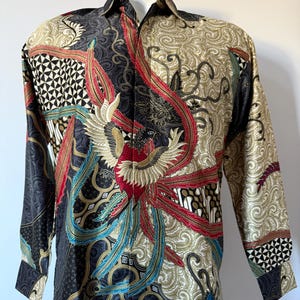 Pode incluir: Uma camisa de manga comprida com botões e uma estampa detalhada em estilo batik. O design apresenta um motivo central de fénix em vermelho, dourado e preto, rodeado por padrões intrincados, formas geométricas e elementos florais em azul e creme.