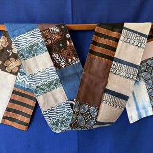 Peut inclure: Un long chemin de table rectangulaire composé d'un patchwork de divers motifs textiles. La pièce présente des sections de tissu marron, bleu, beige et noir avec des imprimés floraux, géométriques, rayés et de style batik disposés en séquence.