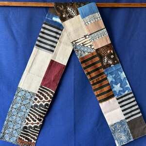 Peut inclure: Une longue écharpe rectangulaire en patchwork composée de divers carrés de tissu présentant des motifs variés, notamment des rayures noires et blanches, des étoiles bleues, des motifs floraux bruns, des formes géométriques et des panneaux unis.