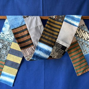 Peut inclure: Un long chemin de table rectangulaire composé d'un assemblage de divers motifs textiles. La pièce présente des sections de tissu bleu teint par nouage, des rayures horizontales aux tons terre, des panneaux gris clair unis et des imprimés noirs et blancs.