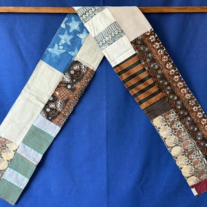 May include: A long rectangular scarf constructed from various fabric patches. The design features a mix of blue star-patterned fabric, solid cream panels, and several sections of brown, black, and tan textiles with intricate geometric and floral prints.