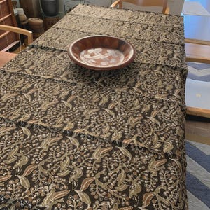 May include: A brown and beige patterned tablecloth with a wooden bowl on top. The tablecloth has a repeating botanical design in shades of brown, beige, and green. The wooden bowl has a lighter-colored floral design.