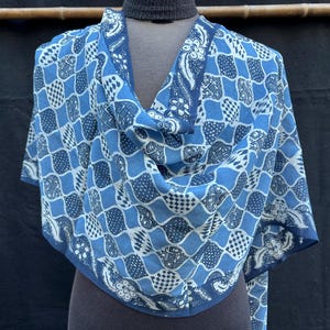 Peut inclure: Écharpe à motifs bleus et blancs drapée sur un mannequin. L'écharpe présente un motif répétitif de carrés et de motifs floraux dans des tons de bleu et de blanc. L'écharpe est en tissu léger.