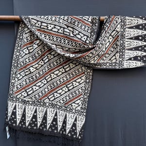 Peut inclure: Un textile rectangulaire présentant un motif batik noir, blanc et rouille. Le design comprend des bandes diagonales de motifs floraux et géométriques, bordées par une rangée de formes triangulaires aux motifs blancs complexes et franges.