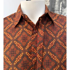 May include: A button-down shirt featuring a repeating geometric pattern in shades of rust orange, dark brown, and golden yellow. The garment has a pointed collar, a front button placket with matching dark buttons, and a single chest pocket.