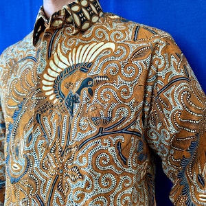 Puede incluir: Una camisa de manga larga con botones que presenta un patrón batik en marrón y crema con intrincados motivos de remolinos y un diseño de ave estilizada. La camisa tiene un cuello en punta y un bolsillo frontal con un detallado estampado textil.