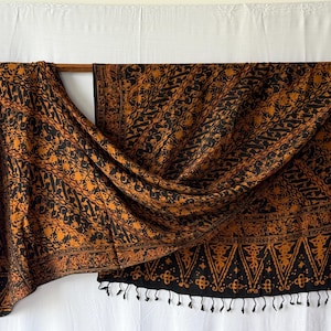 Peut inclure: Un textile rectangulaire présentant un motif batik noir et orange brûlé avec des motifs floraux et géométriques complexes. Le tissu affiche un design répétitif sur toute la surface et est fini avec une rangée de petites franges noires sur le bord inférieur.