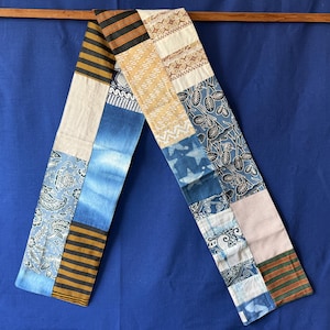 Peut inclure: Une longue écharpe rectangulaire en patchwork composée de divers segments de tissu. Le textile présente un mélange de motifs incluant des imprimés floraux bleu et blanc, des motifs batik géométriques beige et marron, et des rayures horizontales.