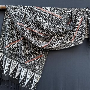 Peut inclure: Un tissu batik noir et blanc orné de motifs tourbillonnants complexes et de bordures géométriques. Le textile présente de fines lignes diagonales aux accents couleur rouille et se termine par une bordure à franges, drapé sur une tige en bois.