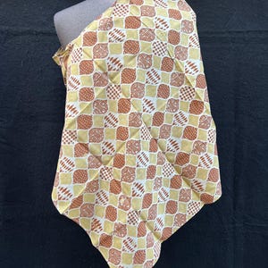 以下が含まれることがあります： マネキンに掛けられた模様入りの生地。生地は、黄色の背景に茶色と白の形が繰り返されるデザインです。生地は、衣類や室内装飾に使用される可能性があります。