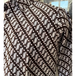 Pode incluir: Uma camisa de manga comprida com botões e um padrão batik diagonal em castanho escuro e branco. A camisa tem um colarinho padrão e um bolso no peito, com um design de tecido composto por formas curvas e repetitivas dispostas em filas.
