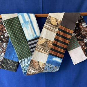 Peut inclure: Un long chemin de table rectangulaire composé d'un assemblage de divers motifs textiles. La pièce présente des sections de tissu noir à motifs floraux blancs, des rayures vertes et dorées, du tie-dye bleu et du tissu beige à motifs bruns.