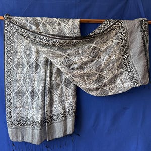 Peut inclure: Une écharpe rectangulaire présentant un motif géométrique noir et blanc avec des motifs floraux complexes. Le tissu affiche un design en losanges répétitif, une large bordure décorative et des franges fines sur le bord inférieur.