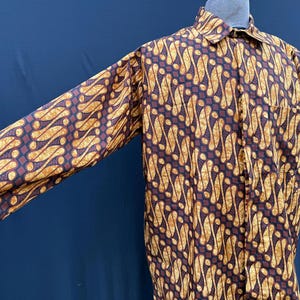 Pode incluir: Uma camisa de manga comprida com botões e um padrão batik marrom e dourado. A camisa tem uma gola clássica e fechamento com botões.