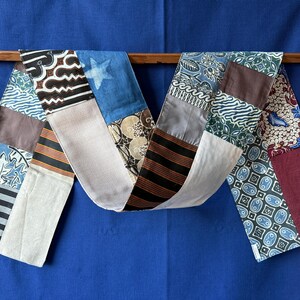 Peut inclure: Une longue écharpe rectangulaire en patchwork composée de divers panneaux de tissu. Les segments textiles présentent des motifs variés, notamment des rayures noires et blanches, des motifs floraux bleus et blancs, des blocs unis marron.