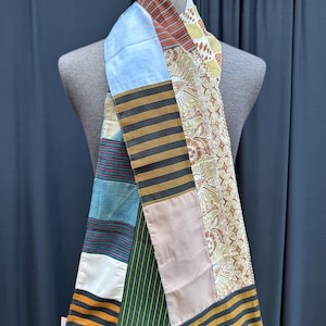 May include: A long, patchwork scarf made of different fabrics. The scarf is made of a variety of colours and patterns, including blue, brown, green, and yellow. The scarf is tied around a mannequin's neck.