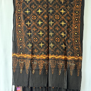 Puede incluir: Un pañuelo rectangular negro con un patrón repetitivo de motivos en forma de estrella amarillos y naranjas dentro de una cuadrícula de líneas curvas. Los bordes tienen un borde geométrico y una línea ondulada, con flecos negros abajo.