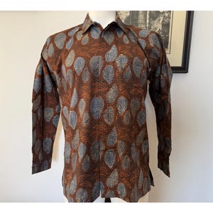 Può includere: Una camicia a maniche lunghe con sfondo marrone e un motivo ripetuto di foglie color azzurro e bianco. La camicia presenta un colletto standard, un'abbottonatura frontale e maniche lunghe con polsini ai polsi.