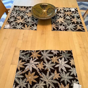 Peut inclure: Une table à manger en bois avec quatre sets de table à motifs de feuilles, un chemin de table assorti et un bol décoratif. Les sets de table et le chemin de table présentent un fond noir avec des motifs de feuilles blanches, beiges et dorées.