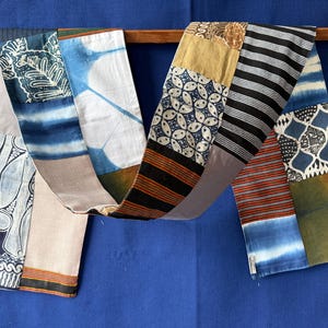 Peut inclure: Une longue écharpe rectangulaire en patchwork composée de divers panneaux de tissu. Le textile présente des motifs variés, notamment du batik bleu et blanc, du tie-dye indigo, des rayures noires et blanches et des tons terreux, assemblés en une bande.
