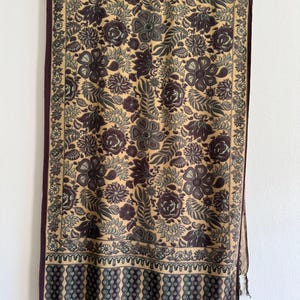 May include: A brown and beige shawl with a floral pattern. The shawl has a fringe border and a dark brown border with a geometric pattern.