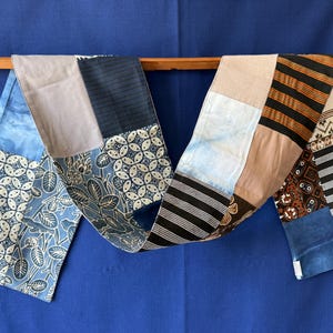 Peut inclure: Une longue écharpe en patchwork composée de divers carrés de tissu dans des tons bleu, beige, noir et marron. Les panneaux textiles présentent des motifs variés, notamment des imprimés batik floraux, des rayures horizontales et géométriques.