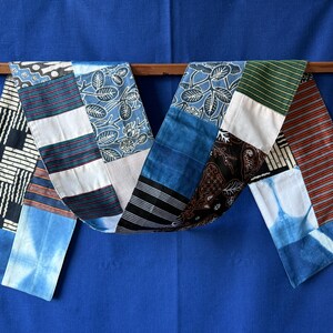 Peut inclure: Une longue écharpe en patchwork rectangulaire composée de divers motifs de tissus, notamment des imprimés floraux bleu et blanc, des designs géométriques noir et blanc, des rayures rouges et bleues, des sections bleu uni et des motifs floraux batik.