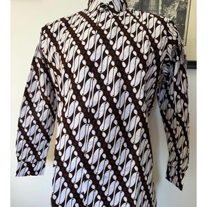 May include: A long-sleeved button-down shirt featuring a dark brown and white batik pattern. The garment has a standard collar, a front button placket, and cuffs on the sleeves. The fabric displays a repeating diagonal motif of curved shapes.