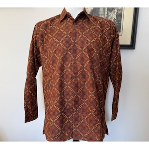 May include: A long-sleeve button-down shirt in a deep brown hue, featuring a repeating geometric pattern of diamonds and small orange and yellow floral-like motifs across the entire fabric, including a standard collar and buttoned cuffs.