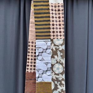 May include: A long, narrow patchwork scarf made of different patterned fabrics. The scarf is made of brown, white, black, and beige fabrics with geometric and floral patterns. The scarf is hanging from a wooden rod.
