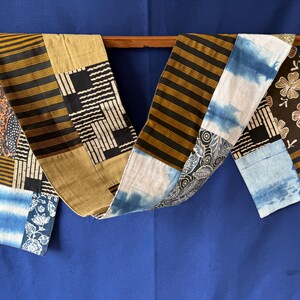 Peut inclure: Un long chemin de table rectangulaire présentant un motif patchwork composé de divers textiles. La pièce comprend des sections à rayures horizontales noires et dorées, des motifs tie-dye bleu et blanc et du tissu brun foncé à motifs floraux blancs.