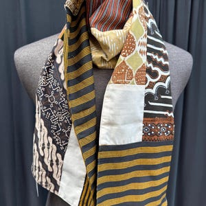 May include: A long, rectangular scarf made from patchwork fabric. The scarf is made of different patterns of black, white, brown, and gold fabric. The scarf is folded in half and draped over a mannequin.
