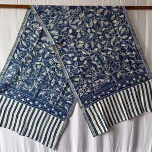Peut inclure: Une écharpe rectangulaire présentant un fond bleu profond avec un motif botanique blanc composé de feuilles et de petits fruits. Les extrémités de l'écharpe sont ornées d'une bande horizontale de petites fleurs suivie de rayures verticales blanches.
