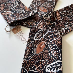 May include: Set of three brown and black patterned fabric napkins with white accents. The napkins feature a leaf and circle design. The napkins are folded and stacked on a white surface. A small tag with the text "The Language of Cloth" is visible in the image.