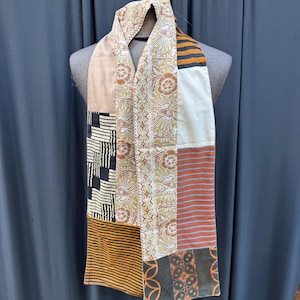 Peut inclure: Une longue écharpe en patchwork avec une variété de motifs bruns, noirs, blancs et oranges. L'écharpe est faite de différents tissus et a un aspect unique et fait main.