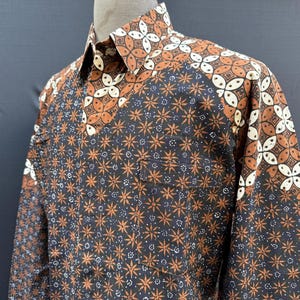 May include: A long-sleeved button-down shirt with a dark navy background and a repeating pattern of orange and white floral and geometric shapes. The collar and upper sleeves feature a larger, more intricate design. The shirt has a single chest pocket.