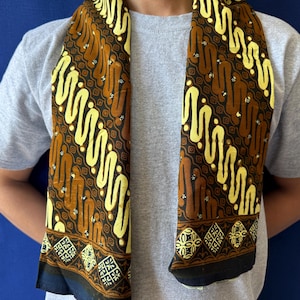 Peut inclure: Une écharpe rectangulaire présentant un motif batik répétitif dans des tons de brun, jaune et noir. Le design se compose de lignes ondulées diagonales et de petits accents floraux, avec une bordure décorative de losanges géométriques.