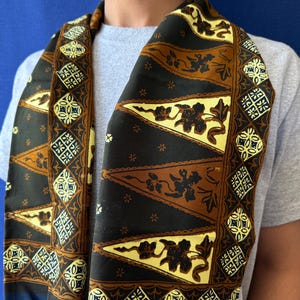 Peut inclure: Une écharpe présentant un motif de style batik avec des éléments triangulaires en noir, marron et jaune pâle. Le design comprend des détails floraux complexes et des formes géométriques en losange disposées en bordure répétitive.