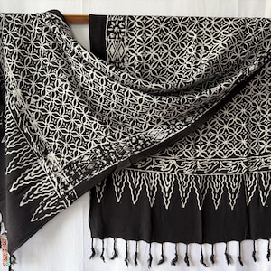 Peut inclure: Un textile noir et blanc présentant un motif géométrique répétitif de cercles et de losanges entrelacés. Le tissu comprend une bordure avec un motif stylisé de flammes ou de feuilles sur le bord inférieur et se termine par des franges noires.