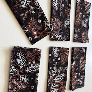 以下が含まれることがあります： 茶色と白の バティック模様が施された黒い布製のナプキンが6枚。模様には葉、花、幾何学模様が描かれている。