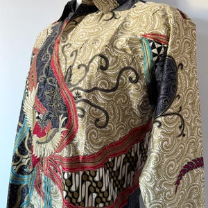 May include: A long-sleeve button-down shirt featuring a complex batik pattern in shades of cream, black, red, and teal. The design includes intricate swirling motifs, geometric shapes, and a prominent bird illustration on the front panel.