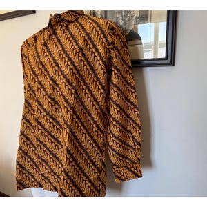 May include: A long-sleeved button-down shirt featuring a repeating diagonal batik pattern in shades of deep brown and golden yellow. The garment has a standard collar, a front button placket, and cuffs at the end of each sleeve with a consistent print.