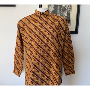 May include: A long-sleeved button-down shirt featuring a repeating diagonal pattern of golden yellow flame-like motifs set against a dark brown background. The shirt has a standard collar, a front button placket, and cuffed sleeves on a mannequin.