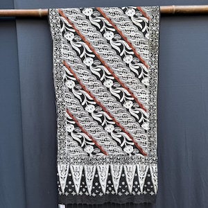 Peut inclure: Un textile rectangulaire présentant un motif batik noir, blanc et terre cuite avec des bandes diagonales de motifs floraux. Le tissu possède une bordure décorative aux formes géométriques et un bord inférieur fini par de petites franges.