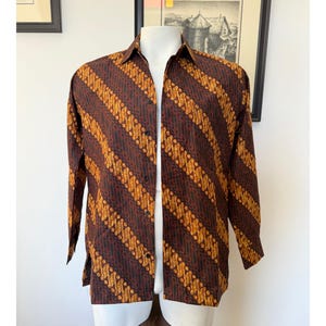 May include: A long-sleeve button-down shirt featuring a dark brown and golden yellow diagonal batik pattern. The shirt has a standard collar, a front button placket with dark buttons, and a straight hemline with side slits, displayed on a mannequin.